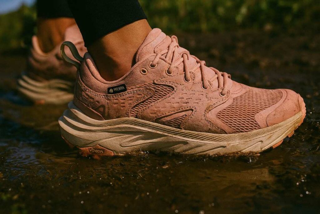 Female hiker testing HOKA Anacapa 2 Low GTX shoes on muddy coastal trail with puddles.