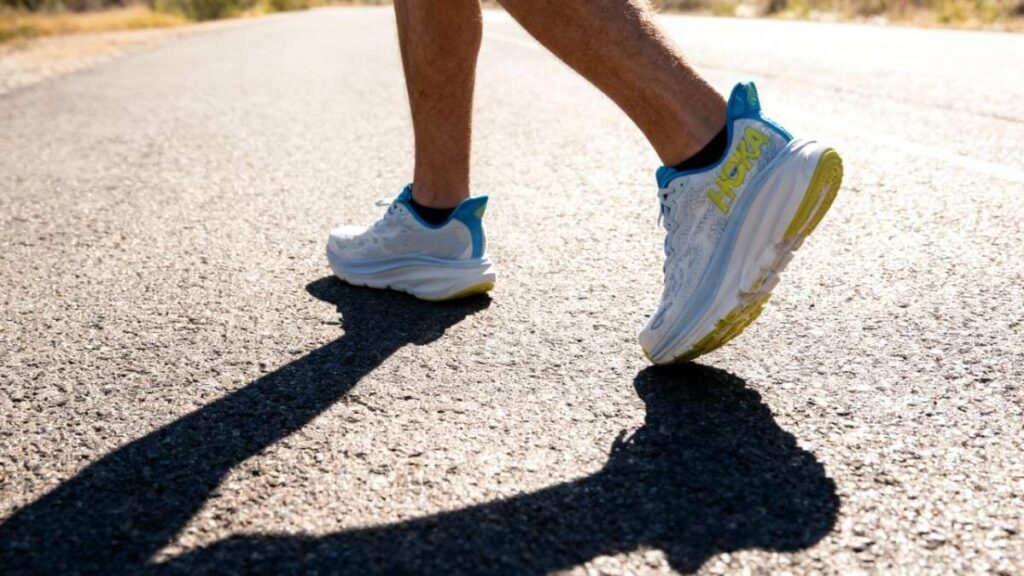 Knees down shot of hiker wearing HOKA Clifton 9 & walking on road.